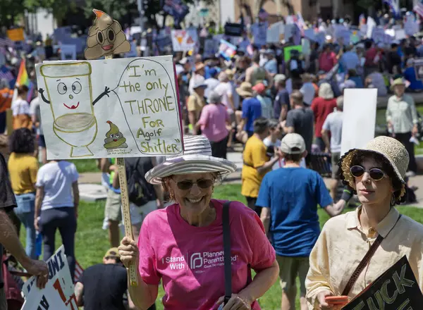 Karikatür tuvaleti ve kaka emojisi olan komik bir tabela tutan bir kadınla protesto sahnesi. Arka plandaki kalabalık, siyasi bir miting veya protesto anlamına gelen çeşitli işaretler taşıyor.