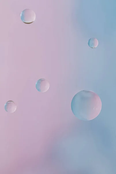 Macro drops of oil on the surface of the water. Delicate cosmetic bubbles background for advertising cosmetic products in soft blue pink tones, copy space and gradient. Vertical photo