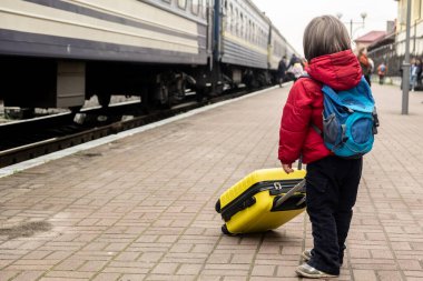  Küçük suratsız çocuk trenin yanındaki platformda duruyor ve elinde sarı bir bavul tutuyor. Tren yolculuğu konsepti