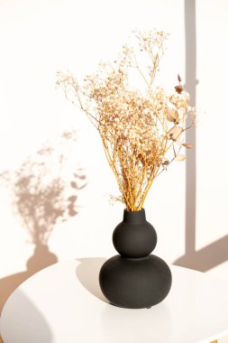 Beyaz masadaki siyah vazoda kurutulmuş çiçek demeti. Güzel gölgeler. İskandinav tarzı iç mekan. Güneşli bir gün