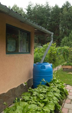 Kırsal bir evin kenarındaki bir yere bağlı mavi yağmur suyu fıçısı, yemyeşil yapraklar ve bahçe yollarıyla çevrili. Yağmur suyu hasat mimarisi. Dikey fotoğraf