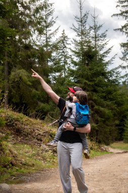 Genç oğlunu taşıyan bir baba yemyeşil bir ormanda yürüyüş yaparken ağaçlara işaret ediyor, açık hava macerasını ve kaynaşmayı vurguluyor. Babalar Günü konsepti 