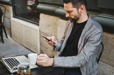 Kafkasyalı bir adam bir sokak kafeteryasında bir masaya oturur ve telefona bakar.