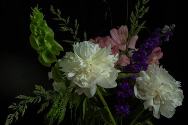 Beyaz şakayık çiçekleri, pembe ve mor çiçekleri ve yeşil yaprakları içeren zarif bir buket, dramatik bir kontrastlık için siyah bir arka planda dizilmiş.