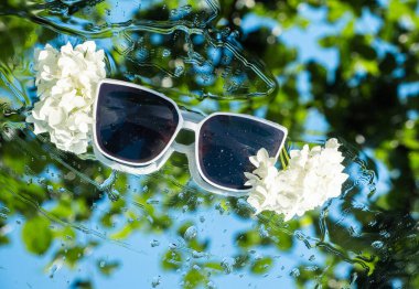 Beyaz güneş gözlükleri, yeşil ve mavi arkaplanlı su yüzeyinde yüzen beyaz çiçeklerle bezenmiş yaz ve rahatlama çağrıştırıyor.. 