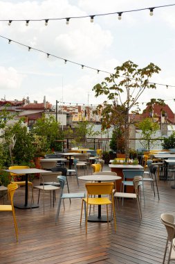 Yeşilliklerle çevrili çeşitli renkli sandalye ve masaların yer aldığı davetkar bir çatı katı kafesi. Açık hava manzarası gökyüzü altındaki şehir manzarası sunuyor.. 