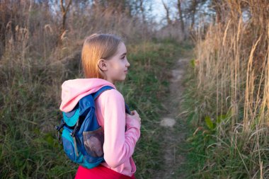  Sırt çantalı beyaz bir kız gün batımında doğa yolunda yürüyor. Pembe bir kapüşonlu giyer, sonbahar sezonunda açık hava ortamının tadını çıkarır..