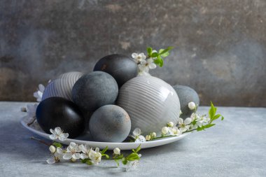 Beyaz bir tabakta, kiraz çiçeği dalıyla tamamlanmış siyah-beyaz dekoratif yumurtaların şık düzenlenmesi. Gelişmiş bir ekran oluşturarak, dokulu bir arkaplana karşı ayarla.