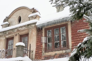 Karla kaplı çatısı, saçaklardan sarkan buz sarkıtları ve kırmızı kiremitli cephesi olan eski bir köy binası. Sakin bir kış sahnesi tarihi mimari ve buz gibi cazibeyi gözler önüne seriyor.. 