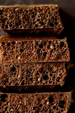 Kahverengi gözenekli çikolata makrosu. Karanlık bir arkaplana karşı üç parça istiflenmiş. Fotoğrafta eşsiz bir desen ve köpüklü çikolatanın derin rengi gösteriliyor. Yumuşak odak.
