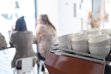 Samimi bir kafede kahve makinesinin üzerinde beyaz bardaklar. Arka planda bulanık insanlar, sıradan atmosfer. Bulanık ve seçici odaklanma. 