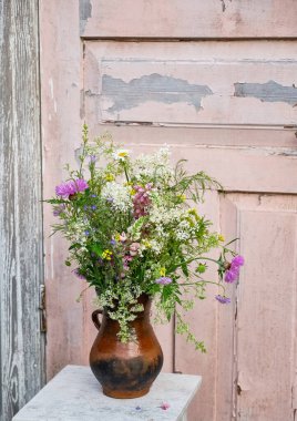 Kır çiçekleri, lupinler ve pembe asterlerin de dahil olduğu yabani çayır çiçekleri, eskimiş ahşap bir masada eski bir kil sürahi içinde eski püskü pembe bir kapıya karşı dizilmiş.. 