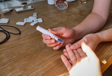 Bir adam bir glukometre tutuyor ve parmağından kan şekerini ölçmeye hazırlanıyor. Sahne, arka planda bir domino oyunu ve tıbbi bir aletle tahta bir masada geçiyor..