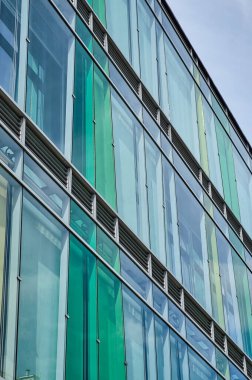 Dikey yeşil ve mavi panellere sahip modern cam cephe, gökyüzünü ve çevreyi minimalist bir mimari tasarımla yansıtıyor. Kentsel geometri