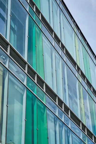 Dikey yeşil ve mavi panellere sahip modern cam cephe, gökyüzünü ve çevreyi minimalist bir mimari tasarımla yansıtıyor. Kentsel geometri