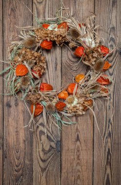 Kurumuş çiçekler, dikenler ve portakallı bir bitki çelengi ahşap bir arka planda rafya ile, sıcak doğal tonlarda, dikey yönelimli, ahşap berrak desenli.