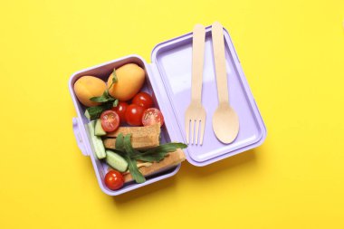 Atıştırmalık, meyve, sebze ve suyla dolu bir beslenme çantasının yanı sıra sarı arka planda okul malzemeleri.