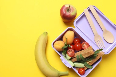 Atıştırmalık, meyve, sebze ve suyla dolu bir beslenme çantasının yanı sıra sarı arka planda okul malzemeleri.
