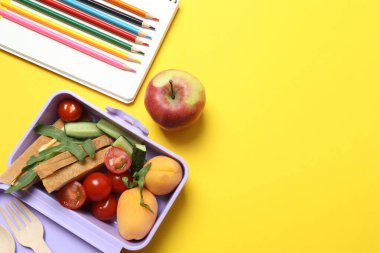 Atıştırmalık, meyve, sebze ve suyla dolu bir beslenme çantasının yanı sıra sarı arka planda okul malzemeleri.