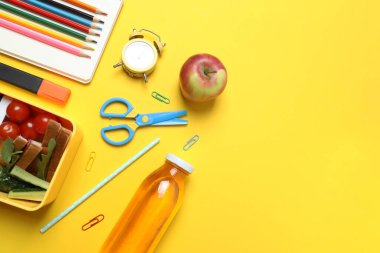 Atıştırmalık, meyve, sebze ve suyla dolu bir beslenme çantasının yanı sıra sarı arka planda okul malzemeleri.