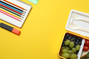 Atıştırmalık, meyve, sebze ve suyla dolu bir beslenme çantasının yanı sıra sarı arka planda okul malzemeleri.