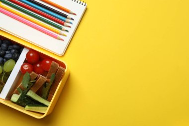 Atıştırmalık, meyve, sebze ve suyla dolu bir beslenme çantasının yanı sıra sarı arka planda okul malzemeleri.