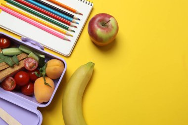 Atıştırmalık, meyve, sebze ve suyla dolu bir beslenme çantasının yanı sıra sarı arka planda okul malzemeleri.