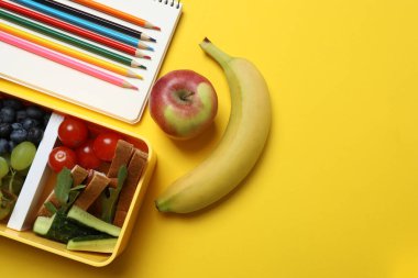 Atıştırmalık, meyve, sebze ve suyla dolu bir beslenme çantasının yanı sıra sarı arka planda okul malzemeleri.