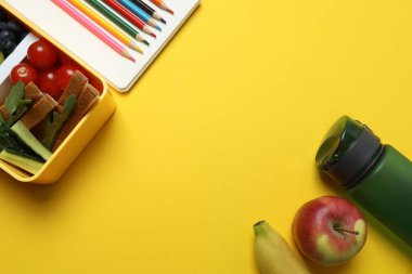 Atıştırmalık, meyve, sebze ve suyla dolu bir beslenme çantasının yanı sıra sarı arka planda okul malzemeleri.