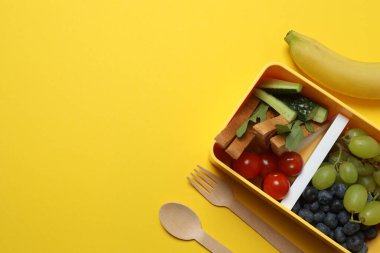 Atıştırmalık, meyve, sebze ve suyla dolu bir beslenme çantasının yanı sıra sarı arka planda okul malzemeleri.