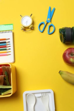 Atıştırmalık, meyve, sebze ve suyla dolu bir beslenme çantasının yanı sıra sarı arka planda okul malzemeleri.