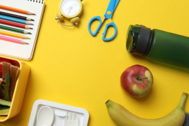 Atıştırmalık, meyve, sebze ve suyla dolu bir beslenme çantasının yanı sıra sarı arka planda okul malzemeleri.