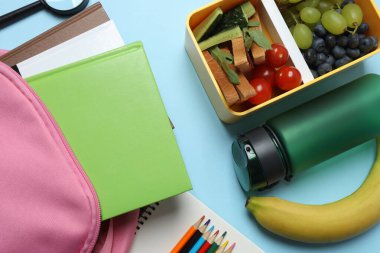 Abur cubur, meyve, sebze ve su ile beslenme çantasının yanı sıra mavi arka planda okul malzemeleri.