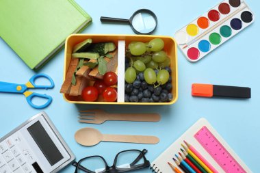 Abur cubur, meyve, sebze ve su ile beslenme çantasının yanı sıra mavi arka planda okul malzemeleri.