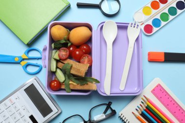 Abur cubur, meyve, sebze ve su ile beslenme çantasının yanı sıra mavi arka planda okul malzemeleri.