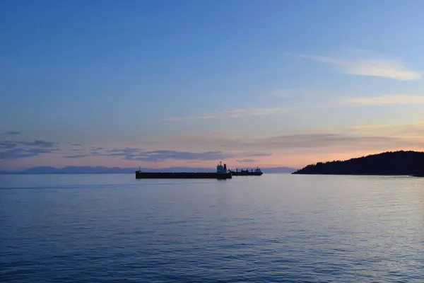 Gün batımında sakin bir deniz manzarası, sakin sularda kargo gemileri, eğimli bir gökyüzü ve uzak dağlarla. Doğa ve deniz aktivitesinin mükemmel bir karışımı.