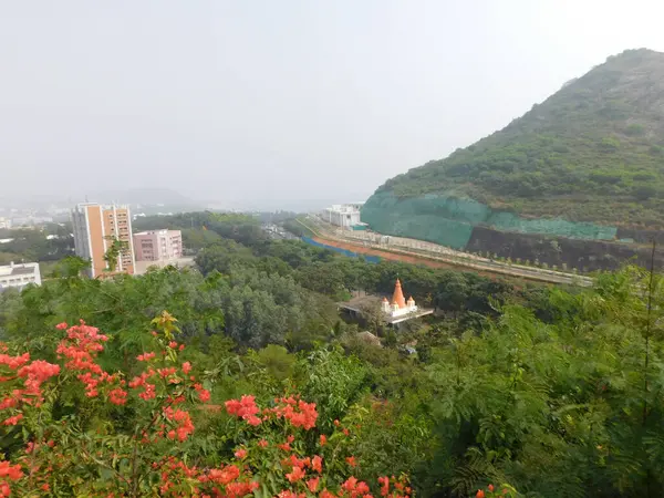  Sağda yeşil bir tepe, orta zeminde modern binalar ve altyapı ve ağaçların arasına yerleştirilmiş küçük bir Hindu tapınağının yer aldığı geniş açılı bir manzara. Ön planda canlı turuncu çiçekler görülebilir..