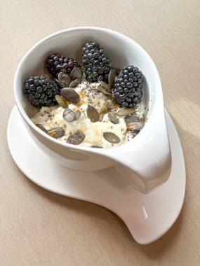 Kabak çekirdeği, chia tohumu, taze böğürtlen ve bal ile kaplı süzme peynirli sağlıklı bir kahvaltı. Güzel beyaz bir seramik bardakta servis edilir. Bir tabağın üzerinde, ahşap bir masanın üzerinde.. 