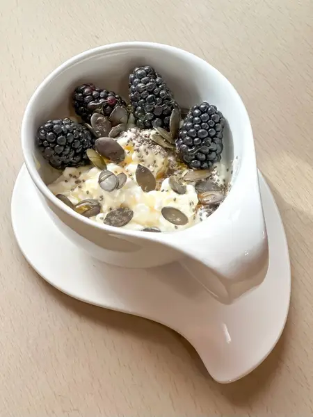 Kabak çekirdeği, chia tohumu, taze böğürtlen ve bal ile kaplı süzme peynirli sağlıklı bir kahvaltı. Güzel beyaz bir seramik bardakta servis edilir. Bir tabağın üzerinde, ahşap bir masanın üzerinde.. 