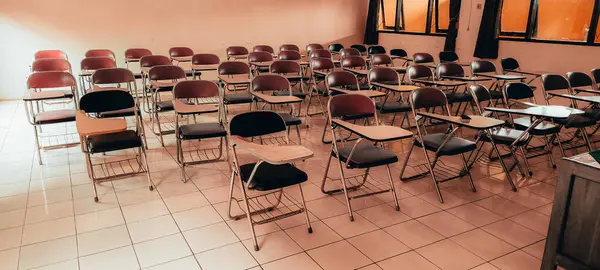 Boş siyah ve kahverengi sandalyeli, yazı masalı, döşemeli ve koyu perdeli pencereli geniş sınıflar..