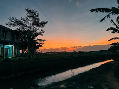 Dar bir kanalda yansıyan turuncu gökyüzü, solda siluetli ev ve ağaçlar ve sağda çerçevelenen muz yaprakları ile huzurlu kırsal günbatımı..