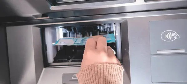 Bir ATM makinesinden mavi banknotları alan bir elin yakın çekimi, sağ tarafında görünür bir temassız ödeme sembolü var.
