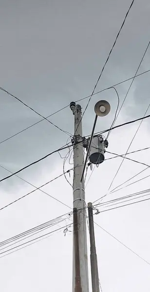 Sokak lambası, trafo ve sayısız elektrik kablosunun yer aldığı bir elektrik direğinin düşük açılı fotoğrafı gri, bulutlu bir gökyüzüne karşı dizilmiş..