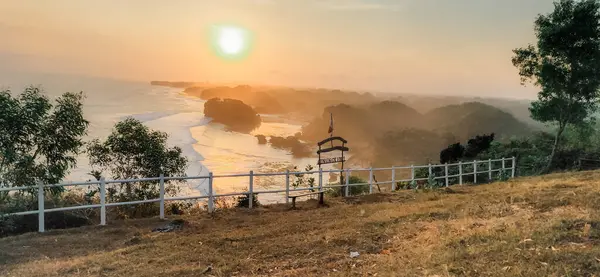 Huzurlu sahil günbatımında beyaz bir çit, çimenli bir tepe, kayalık sahil şeridi ve sakin sahneyi aydınlatan ılık altın bir ışık var..