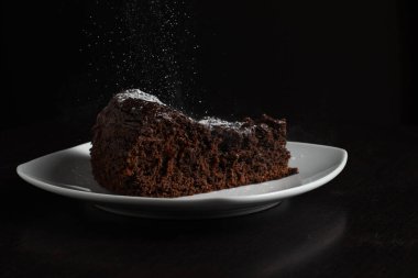 Lezzetli bir dilim çikolatalı kek beyaz bir tabakta siyah bir arka planda zarif bir şekilde sunuluyor. Nemli pasta ve gölgeli çevre arasındaki zıtlık, tatlının zengin ve yoğun dokusunu vurgular..