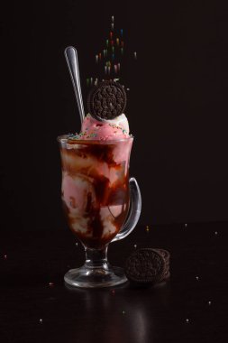 Üstünde çikolatalı kurabiye olan bir bardak dondurma, üzerine sos serpiştirilmiş ve düşen şeker toplarıyla süslenmiş..