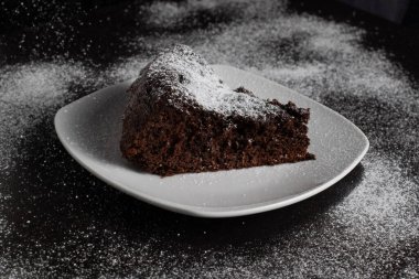 Pudra şekerli çikolatalı pasta dilimi, üzerine pudra şekeri serpilmiş bir masada servis edilir..