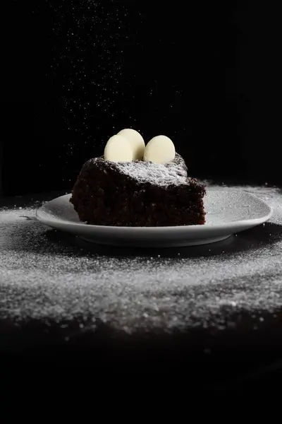 Beyaz bir tabakta servis edilen lezzetli çikolatalı pasta, beyaz çikolata parçacıkları ve toz şekerle süslenmiş..