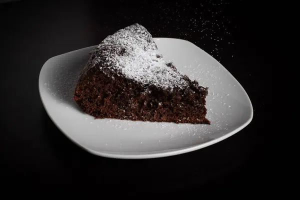Beyaz bir tabakta bir dilim çikolatalı pasta, yukarıdan gelen pudra şekerli, siyah bir arka planda, tatlıya biraz zarafet katıyor..