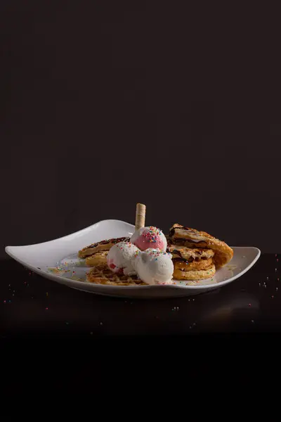 Çilekli ve vanilyalı dondurma ile servis edilen waffle, renkli serpiştirmeler, waffle külahı ve çikolata sosu, koyu arkaplanlı beyaz bir tabakta..